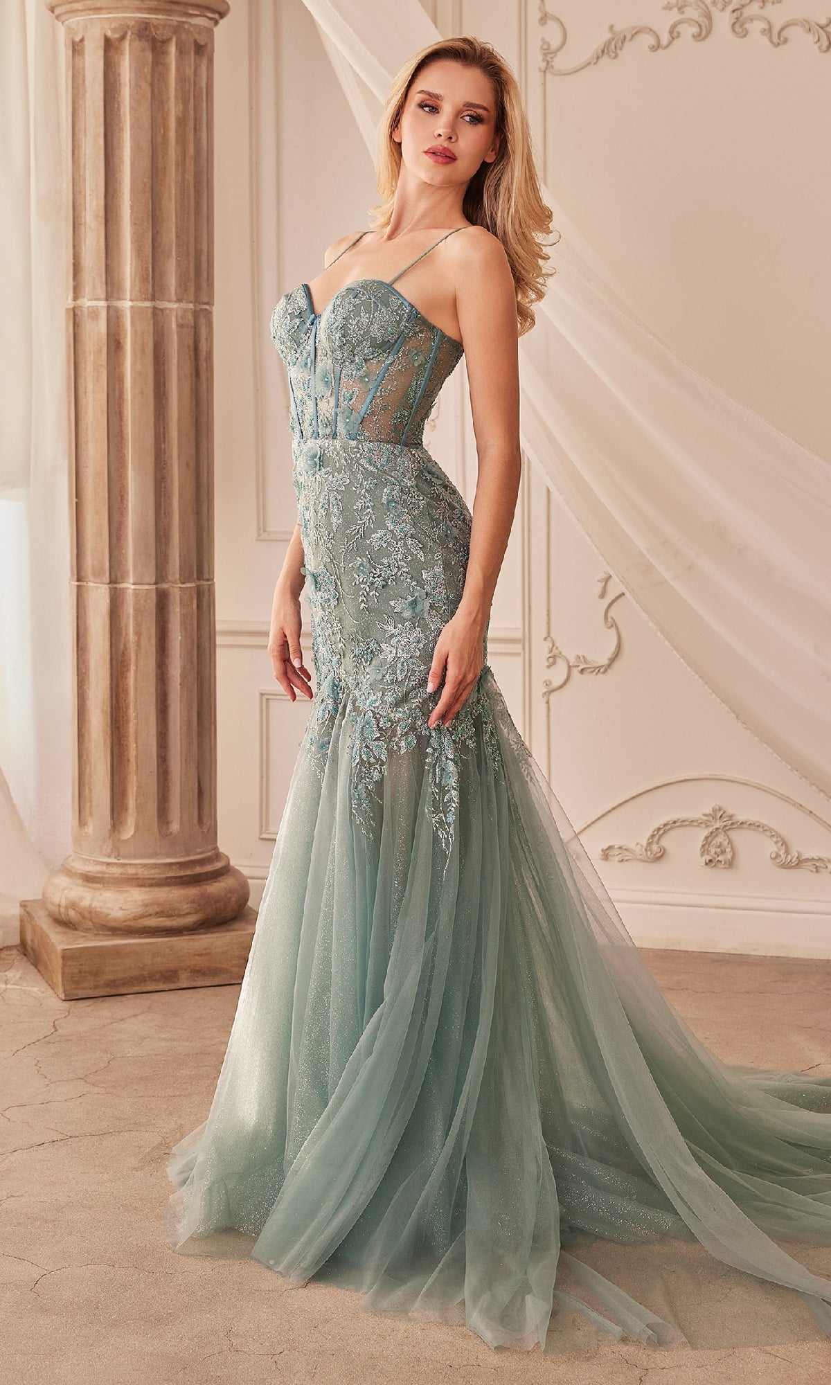 A woman in the Andrea and Leo A1232 long lace mermaid prom dress, featuring a strapless, fitted, pale blue-green bodice and sheer tulle skirt, stands in an elegant classical-style room.
